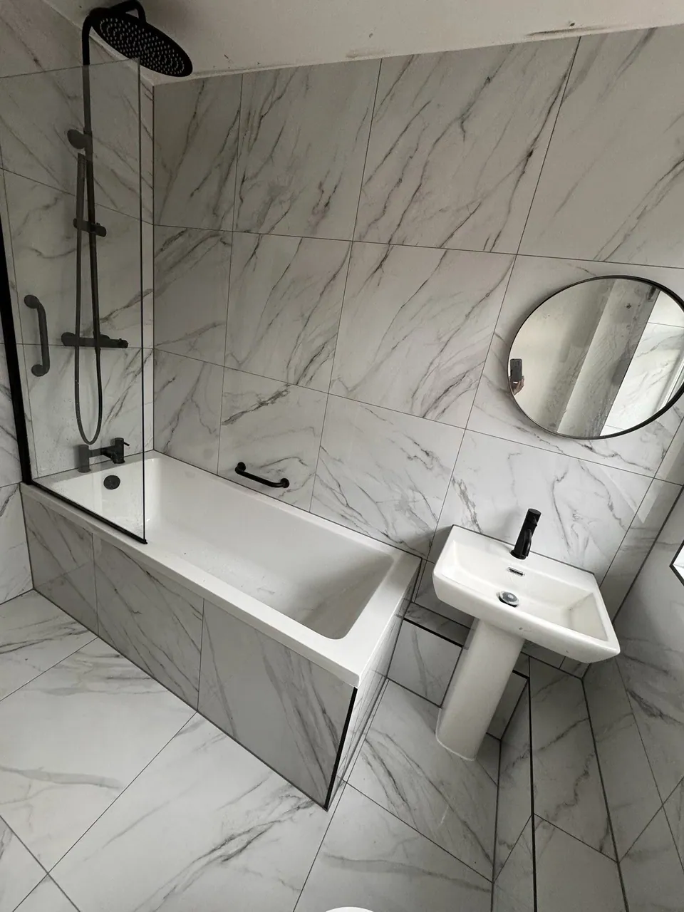 Bathroom in marble-effect tiles with matt black bath screen, black shower and taps and a round black-framed mirror, fitted by Basingstoke Bathrooms